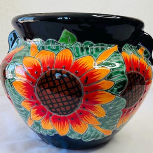 Op de afbeelding: Een grote, ronde keramische bloempot met een glanzende zwarte rand en handvatten. De bloempot is versierd met levendige zonnebloemen in tinten rood, oranje en geel, tegen een groene achtergrond.