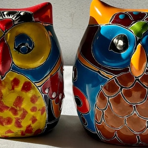 以下が含まれることがあります： 複雑なデザインが施された、カラフルなセラミック製のフクロウの置物2つ。1つのフクロウは、青、オレンジ、茶色で、茶色と黒の模様の腹をしています。もう1つのフクロウは、黄色、赤、青で、黄色と赤の模様の腹をしています。