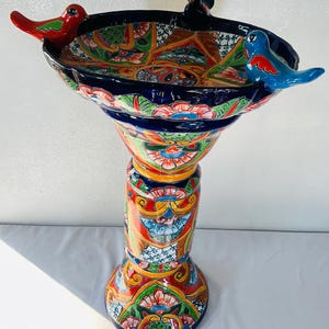 May include: A ceramic birdbath featuring a vibrant, multi-colored hand-painted floral pattern in shades of red, blue, green, yellow, and white. The basin is adorned with three sculpted birds in red, black, and blue perched on the rim.