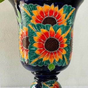 Może przedstawiać: Duży, niebieski ceramiczny pojemnik na rośliny z podstawą w kształcie cokołu. Pojemnik jest ozdobiony ręcznie malowanymi słonecznikami i zielonymi liśćmi.