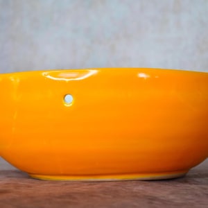 May include: A bright orange ceramic bowl with a small hole in the side. The bowl is round and has a smooth, glossy finish.