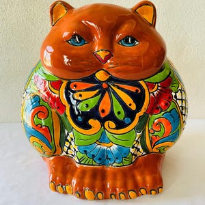 Könnte beinhalten: Keramik-Katzenfigur mit braunem Körper und Kopf, blauen Augen und einem farbenfrohen Blumendesign auf der Brust. Die Figur hat eine glänzende Oberfläche und steht aufrecht.