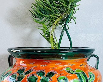 May include: A round, glazed ceramic planter with two side handles, featuring a vibrant orange background decorated with hand-painted green cacti, red accents, and black outlines. A green succulent plant with a single yellow bud grows inside.