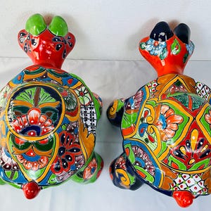 Könnte beinhalten: Zwei Keramik-Schildkrötenfiguren mit lebendigen, handbemalten Designs. Die Schildkröten haben rote Köpfe, grüne und schwarze Akzente und farbenfrohe Panzer mit floralen und geometrischen Mustern in Blau, Orange, Grün und Gelb. Die Figuren sind dekorativ und künstlerisch.