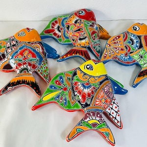 Op de afbeelding: Een verzameling van vier keramische vissen als wanddecoratie, elk voorzien van een levendig, handgeschilderd, veelkleurig bloemen- en geometrisch patroon. De vissen zijn afgewerkt met een glanzend glazuur in oranje, geel, groen en blauw.