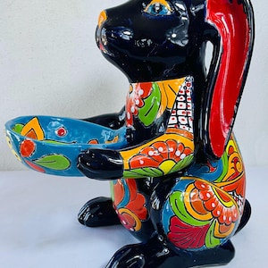 以下が含まれることがあります： 光沢のある黒色のセラミックウサギの置物で、青いボウルを持っています。ウサギはオレンジ、黄色、緑、赤のカラフルな花柄で飾られています。耳の内側は赤色で、目は青色です。