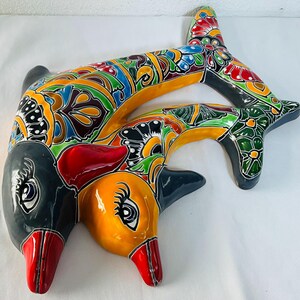Op de afbeelding: Een keramieken wandsculptuur met twee in elkaar verstrengelde dolfijnen, versierd met een levendig, veelkleurig bloemen- en geometrisch patroon in geel, rood, groen, blauw en zwart, met glanzende, handgeschilderde details en duidelijke ogen.