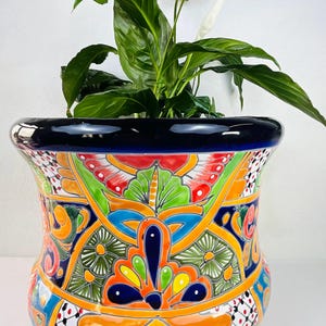 Könnte beinhalten: Ein großer, dekorativer Keramik-Blumentopf mit einem lebendigen, handgemalten Design. Der Topf hat einen schwarzen Rand und ein farbenfrohes Muster aus orangefarbenen, grünen, blauen und roten Blumen- und geometrischen Formen. Er enthält eine grüne Pflanze mit großen Blättern und einer weißen Blüte.