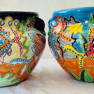 Puede incluir: Dos macetas de cerámica con una vibrante escena submarina pintada. Las macetas presentan peces coloridos, corales y algas sobre un fondo azul y naranja. Cada maceta tiene un borde y asas de color azul oscuro.