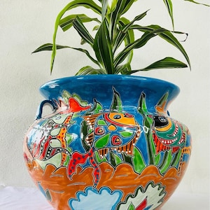 May include: A large, hand-painted terracotta planter with a blue glaze and colorful fish and floral designs. The planter is filled with a green plant.