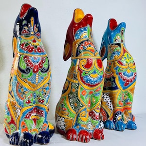May include: Three vibrant ceramic dog sculptures, each with its head raised and mouth open. The dogs are adorned with detailed patterns in red, yellow, blue, green, and orange. The sculptures are displayed on a white surface.