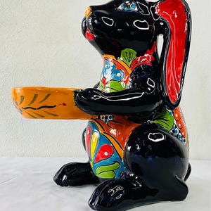 Könnte beinhalten: Eine schwarze Keramik-Hasenfigur mit bunten Blumenmustern. Der Hase hält eine kleine orange Schale in seinen Vorderpfoten.