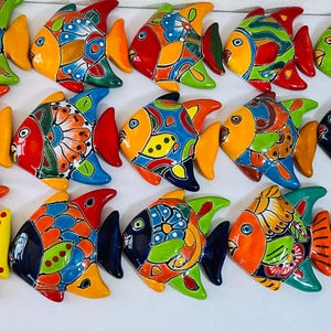 Op de afbeelding: Een verzameling kleurrijke keramische vissculpturen. Elke vis heeft een uniek ontwerp met levendige tinten geel, oranje, rood, blauw en groen. De vissen hebben gedetailleerde patronen en zijn in rijen gerangschikt.