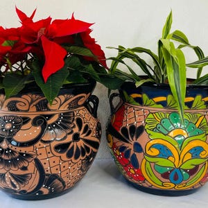 Op de afbeelding: Twee terracotta potten met kleurrijke handgeschilderde ontwerpen. De pot links heeft een zwart-wit bloemenontwerp, terwijl de pot rechts een veelkleurig bloemenontwerp heeft met groene, blauwe, gele en rode accenten.