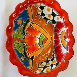 May include: A hand-painted, oval-shaped ceramic dish with a scalloped orange rim. The interior features a vibrant abstract design with orange, red, blue, green, and white colors. The dish is likely for decorative or serving purposes.