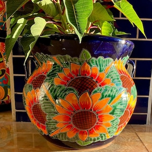 May include: A blue ceramic planter with two handles, decorated with hand-painted sunflowers in shades of yellow, orange, red, and green.