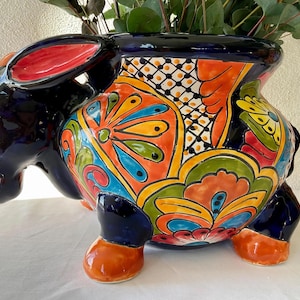 May include: A colourful ceramic donkey planter with a blue base and intricate floral designs in shades of red, orange, yellow, green, and blue. The donkey has a raised head and ears, and its legs are sculpted in a standing position.