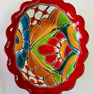 May include: A decorative ceramic bowl with a scalloped red rim. The interior features a vibrant, abstract design with blue, orange, green, and red floral patterns outlined in black. The bowl is likely handmade.