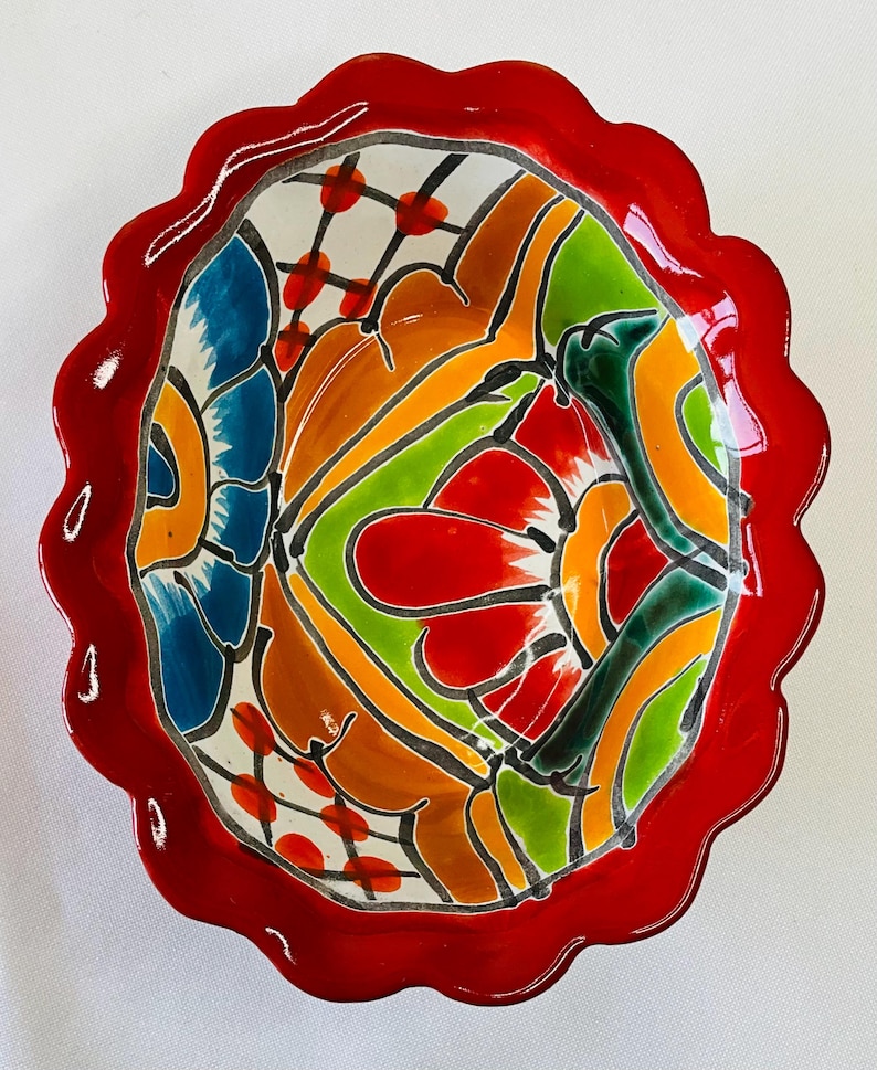 May include: A decorative ceramic bowl with a scalloped red rim. The interior features a vibrant, abstract design with blue, orange, green, and red floral patterns outlined in black. The bowl is likely handmade.