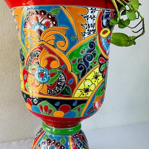May include: A colourful, hand-painted ceramic planter with a pedestal base. The planter has a bold red rim and base, with intricate floral and geometric patterns in blue, orange, green, and yellow. Green leafy plant stems cascade over the rim.
