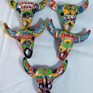 May include: Five colorful ceramic bull skull ornaments. Each skull features a unique hand-painted design with vibrant colors including red, blue, green, and yellow. The horns are curved, and each skull has a loop for hanging.
