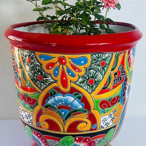 Könnte beinhalten: Ein großer, runder Keramik-Pflanztopf mit glänzend rotem Rand und Fuß, verziert mit einem lebendigen, handbemalten Blumen- und Geometriemuster in Gelb, Blau, Grün und Rot. Im Topf befindet sich ein kleiner Rosenstock mit rosa und gelben Blüten.