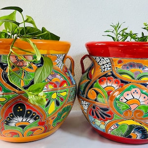 May include: Two brightly coloured ceramic plant pots with detailed floral patterns. One has an orange rim, the other red. Both are filled with greenery, adding a natural element to the decorative pots. Each pot has a pair of handles.
