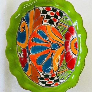 May include: A brightly colored, oval-shaped ceramic bowl with a scalloped green rim. The interior features a vibrant floral design in orange, blue, red, and green. The bowl is likely handmade and suitable for serving or display.