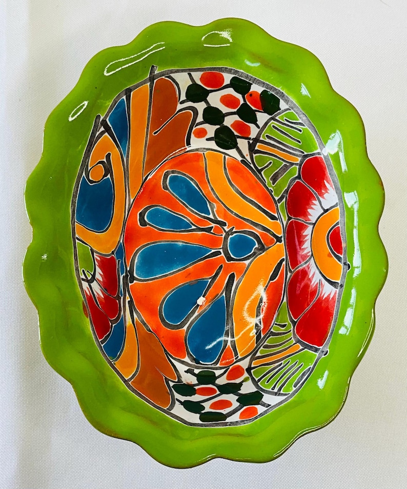 May include: A brightly colored, oval-shaped ceramic bowl with a scalloped green rim. The interior features a vibrant floral design in orange, blue, red, and green. The bowl is likely handmade and suitable for serving or display.