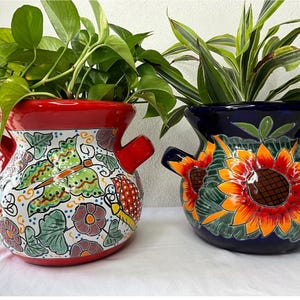 Peut inclure: Deux pots en terre cuite peints à la main avec des motifs floraux colorés. Le pot de gauche est rouge avec un motif de papillon et le pot de droite est bleu avec un motif de tournesol.