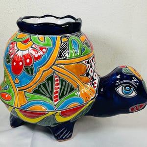 以下が含まれることがあります： 鮮やかな手描きのデザインが施された、カメの形をしたセラミックプランター。カメの体には、赤、青、オレンジ、緑のカラフルな花柄が施されています。頭と脚は濃い青色で、プランターにはスカラップの縁が付いています。