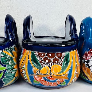 May include: Three hand-painted ceramic planters with a unique kettlebell shape. Each planter features a vibrant design with a dark blue base, a white interior, and colorful floral patterns in orange, yellow, and green. The planters have a handle.