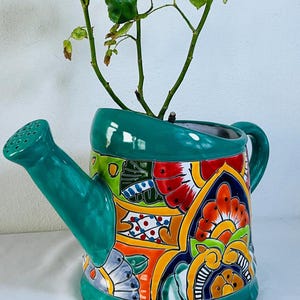 Puede incluir: Una regadera de cerámica utilizada como maceta en un acabado verde azulado vibrante, con un patrón pintado a mano estilo Talavera con motivos florales en rojo, naranja, amarillo y azul. Una pequeña planta verde crece desde la abertura.