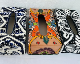 May include: Three rectangular tissue box covers with decorative designs. One is navy blue and white, another is tan with orange and green floral patterns, and the third is black and white. Each has an oval opening.