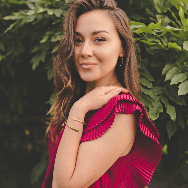 May include: A woman with long brown hair wearing a red pleated top with a gold bracelet on her wrist. She is standing in front of a green leafy background.