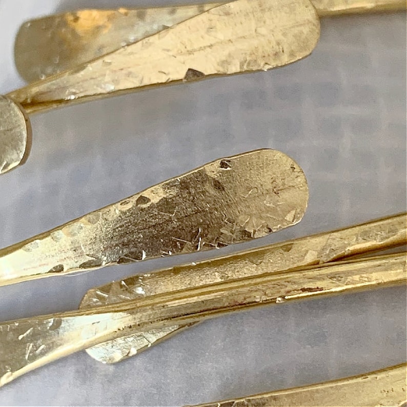 May include: Close-up of a group of gold-colored metal pieces, each with a hammered texture and a slightly rounded, pointed end. The pieces are arranged in a random pattern, creating a visually interesting and textured composition.
