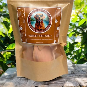 May include: A brown paper bag of dog treats labeled "Back Country Barks Sweet Potato." The label features a dog wearing sunglasses and the text "Dog Gear Company Founded in 2025." The bag contains dehydrated sweet potato treats, net weight 4 oz (113g).