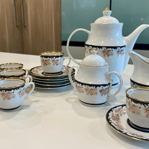 May include: A white porcelain tea set with black and gold accents and floral designs. The set includes a teapot, creamer, sugar bowl, and six cups and saucers.