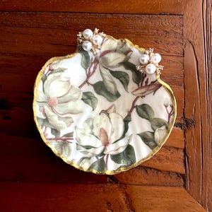 May include: A decorative seashell-shaped dish with a floral magnolia design and gold trim. Two pearl and rhinestone earrings rest on the dish. The dish is on a dark wooden surface.