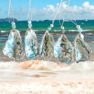 May include: Five hanging oyster shell ornaments with silver string and bead accents. Each shell is decorated with a blue and green aquatic design featuring seaweed and starfish. The shells are suspended above a sandy beach with ocean waves in the background.