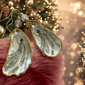 May include: Two gold-trimmed oyster shell ornaments with painted angel figures inside. The ornaments are hanging from a string with white and gold beads.