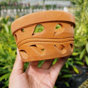 May include: A round terracotta plant pot featuring a series of teardrop-shaped cutouts arranged in two horizontal rows, separated by a raised wavy line that encircles the exterior of the clay vessel.