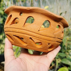 May include: A small, unglazed terracotta planter with a series of decorative cut-out shapes. The oval-shaped pot has a wide rim and is held in a hand. The natural clay color is visible throughout the planter.