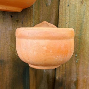 May include: A terracotta-colored, wall-mounted planter with a rounded bowl shape. The planter has a small, decorative element at the top for hanging. The background is a weathered wooden surface.