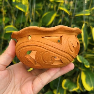 May include: A small, terracotta-colored ceramic pot with a wavy rim and carved floral designs. The pot has several cut-out shapes and is held in a hand against a backdrop of green foliage. Ideal for small plants.