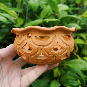 May include: A small, terracotta-colored ceramic pot with a scalloped rim and intricate cut-out designs. The pot is round with a wide opening, perfect for small plants or decorative use. The background is a blur of green foliage.
