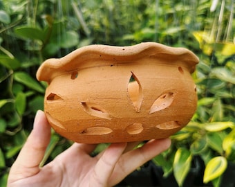 May include: A small, unglazed terracotta pot with a scalloped rim and decorative cut-out leaf shapes. The pot is held in a hand against a backdrop of green foliage, showcasing its natural, earthy tones. Ideal for small plants or succulents.