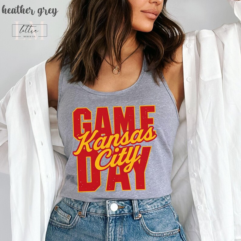 May include: Heather grey tank top with the text "GAME Kansas City DAY" in red and yellow. The tank top is worn with blue jeans and a white button-down shirt. The top is from the brand "lottie merch co."