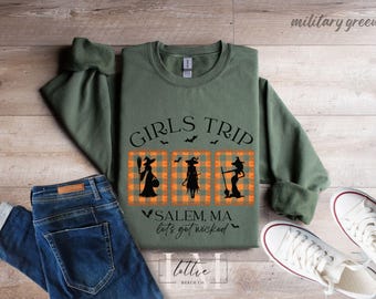 May include: Military green sweatshirt with the text "GIRLS TRIP" and three witch silhouettes on a plaid background. The text "SALEM, MA lets get wicked" is also visible. The sweatshirt is paired with blue jeans and white sneakers.