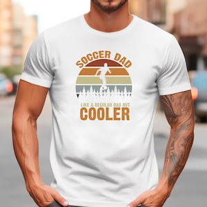 May include: White t-shirt with a retro design featuring the text "SOCCER DAD" above an illustration of a soccer player kicking a ball. The text "LIKE A REGULAR DAD BUT COOLER" is below the illustration.